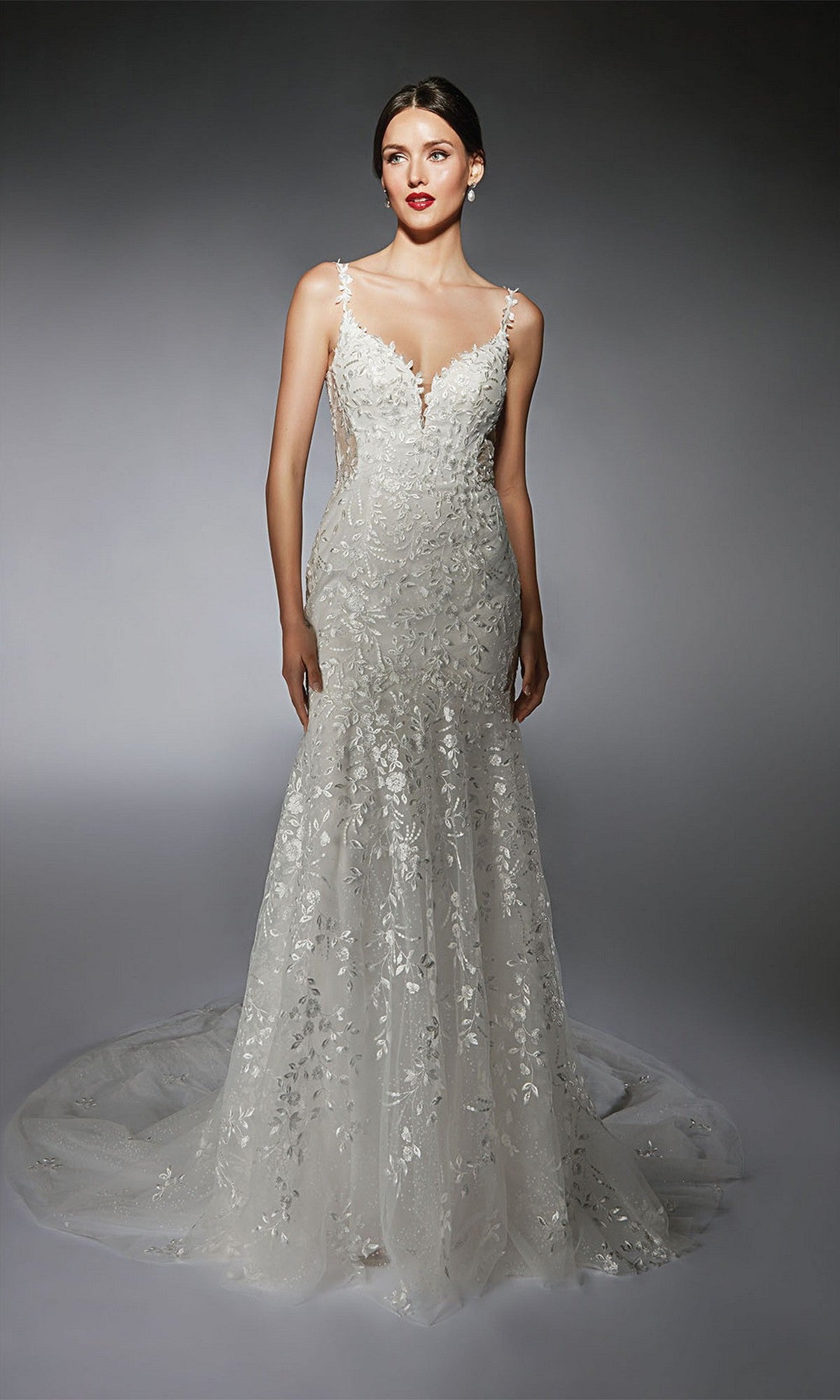 A woman models the Alyce 7122 long white dress, a fitted, sleeveless gown featuring floral lace appliqués and a sweeping train, against a gray background.